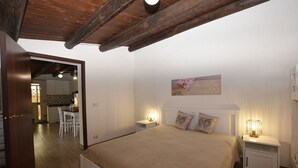 A bedroom with a bed, bedside tables, a nightstand, a wall-mounted picture, and a door leading to a kitchen. - Villa Cigno Reale by Geco Vacation Rentals (Chiaramonte Gulfi)