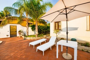 Eine Terrasse mit weißen Liegestühlen, einem großen Sonnenschirm und einer Steinmauer mit Bepflanzung.