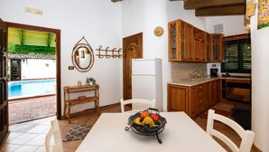 A kitchen with wooden cabinets, a white refrigerator, and a dining table with chairs. - Villa Cigno Reale by Geco Vacation Rentals (Chiaramonte Gulfi)