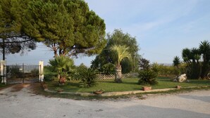 A gravel driveway leading to a gated entrance, surrounded by a variety of trees and potted plants. - Villa Cigno Reale by Geco Vacation Rentals (Chiaramonte Gulfi)