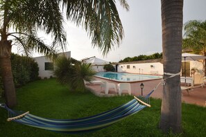 Un espace piscine agrémenté d’un hamac, de palmiers et d’un bâtiment blanc doté d’un toit en tuiles.