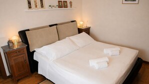 A neatly made bed with white linens and two rolled towels, a wooden nightstand with a lamp, and a shelf with framed pictures and small plants. - Grandparents' Cottage, 15 min from Lake Garda, View of Monte Baldo, Free Parking (San Zeno)