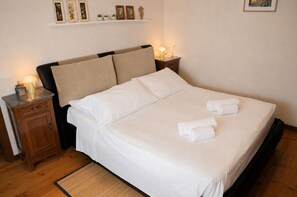 Ein ordentlich bezogenes Bett mit weißen Leinen und zwei aufgerollten Handtüchern, ein Nachttisch aus Holz mit einer Lampe und eine Ablage mit gerahmten Bildern und kleinen Pflanzen.