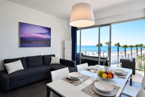 Ein modernes Zimmer mit Esstisch für zwei Personen, einer Couch, einem großen Fenster mit Blick auf den Strand und einem gerahmten Bild an der Wand.