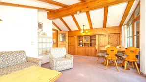 Ein gemütliches Zimmer mit Holzbalkendecke, einer Couch, einem Sessel, einem Esstisch mit Stühlen und eingebauten Holzschränken.