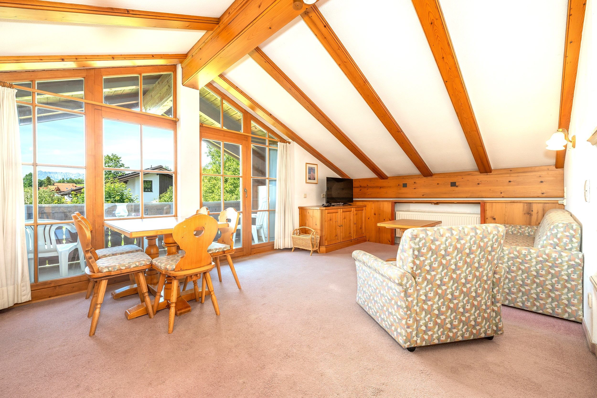 A room with a dining table and chairs, a sofa, a television, and large windows offering a view of the outdoors.
