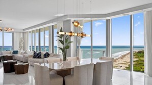 A modern living room with a large glass table and chairs, a sofa set, and a view of the ocean.