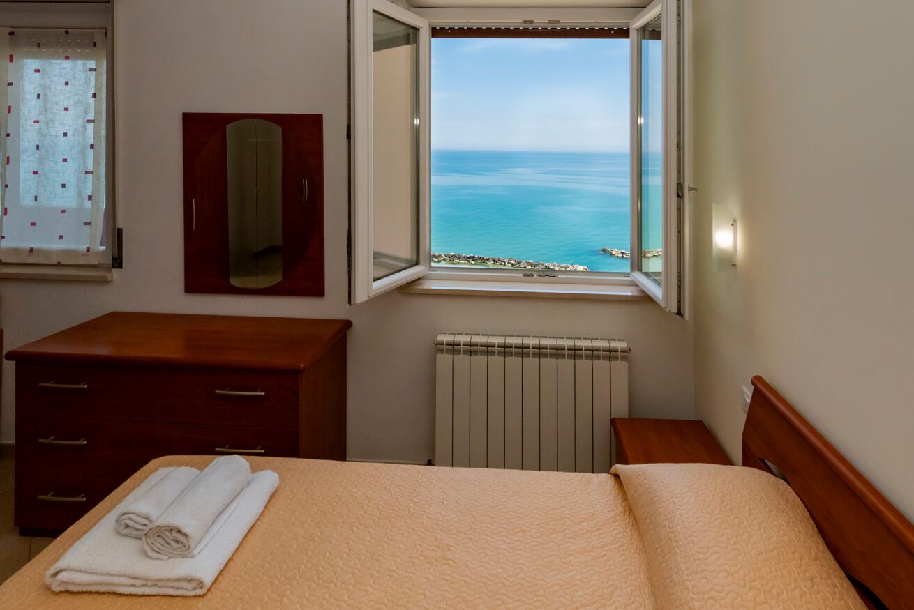 A hotel room with a bed, a wooden dresser, a mirror, a window with a view of the sea, and a radiator.