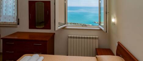 A hotel room with a bed, a wooden dresser, a mirror, a window with a view of the sea, and a radiator.