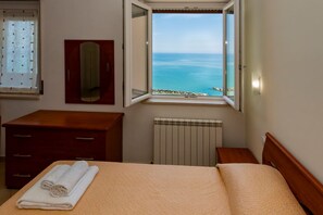 A hotel room with a bed, a wooden dresser, a mirror, a window with a view of the sea, and a radiator. - GuestHost - Sea View Moro Apartment (Montesilvano)
