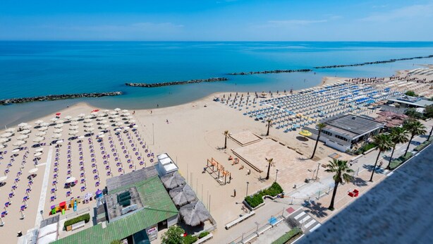 Ein Strand mit Reihen von Sonnenschirmen und Liegestühlen, ein Gebäude mit grüner Dachbegrünung und einem klaren, blauen Himmel.