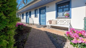 Een rij blauwe deuren met het nummer '73a' op het gebouw, een bank met kussens en roze bloemen in de tuin.