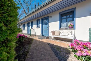 Eine Reihe blauer Türen mit der Nummer '73a' an der Fassade, eine Bank mit Kissen und pinkfarbene Blumen im Garten.