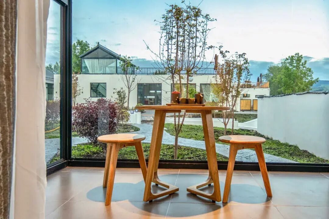 A modern room with a large window, a wooden table with two stools, and a view of a residential area.