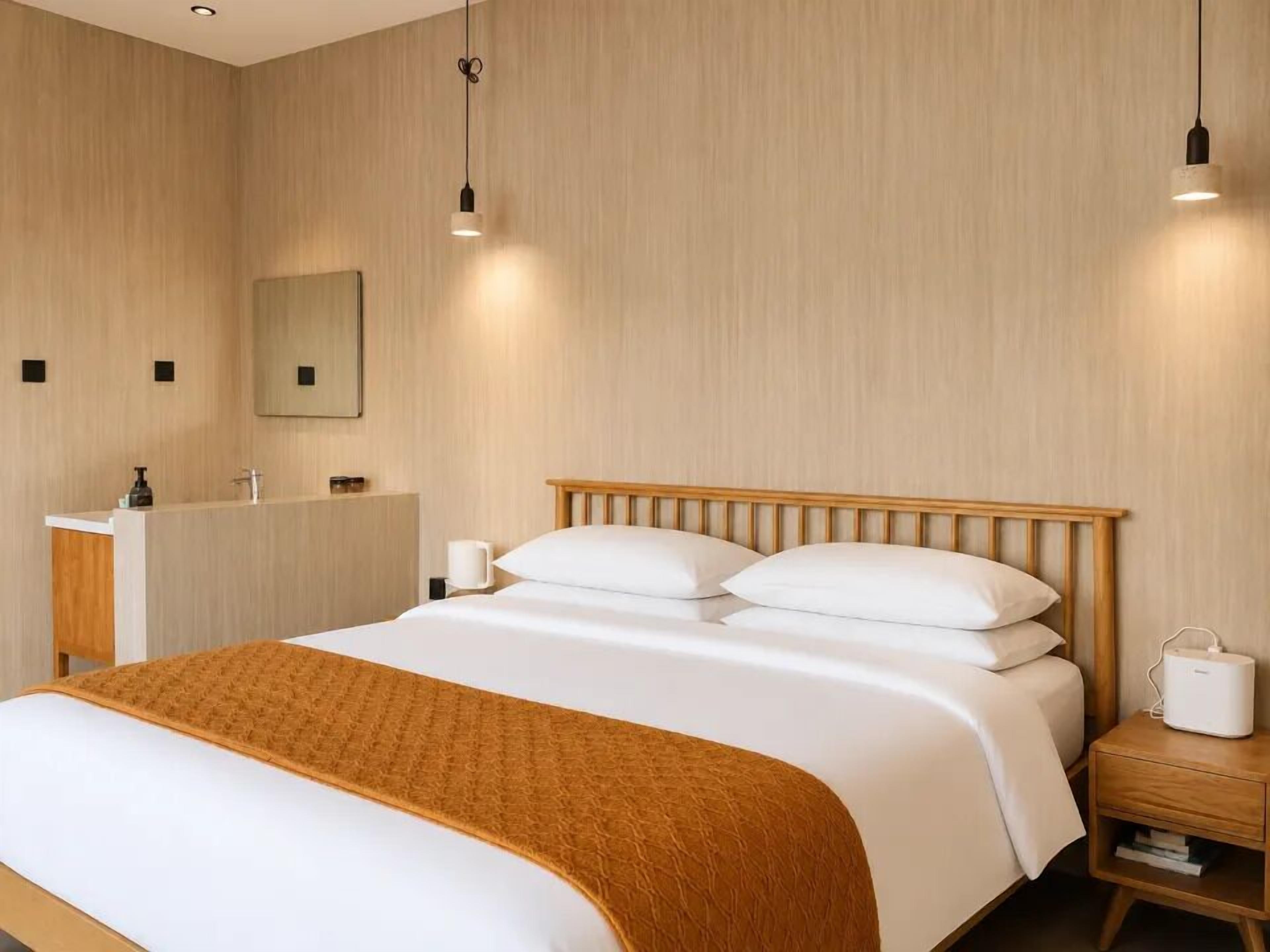 Habitación de hotel con una cama de cabecera de madera, una mesita de noche con lámpara, un espejo y otra mesita más pequeña con un dispositivo.