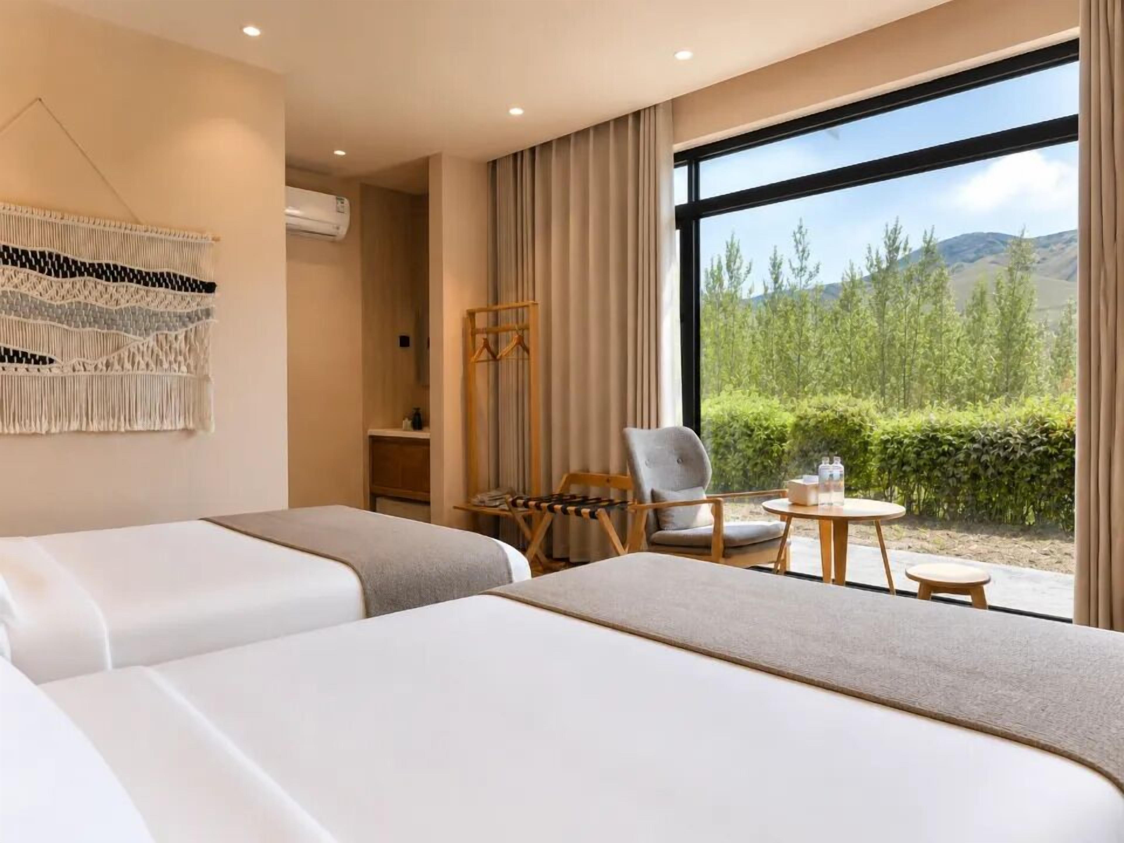 A hotel room with two beds, a chair, a small table, a macrame wall hanging, and a large window with a view of greenery and hills.