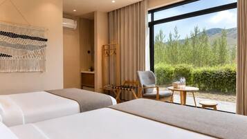 A hotel room with two beds, a chair, a small table, a macrame wall hanging, and a large window with a view of greenery and hills.