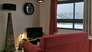 En stue med en rød sofa, et sort tv-bord, et ur, en lampe og et vindue med udsigt.