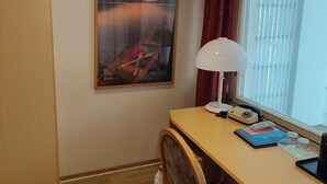 Una habitación con escritorio de madera, una silla, una lámpara, un ventanal con cortinas y un cuadro en la pared.