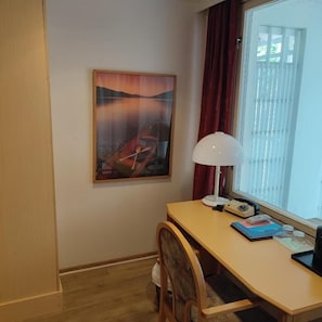 Huoneessa on puinen työpöytä, tuoli, lamppu, ikkuna, jossa on verhot, ja seinällä on taulu.