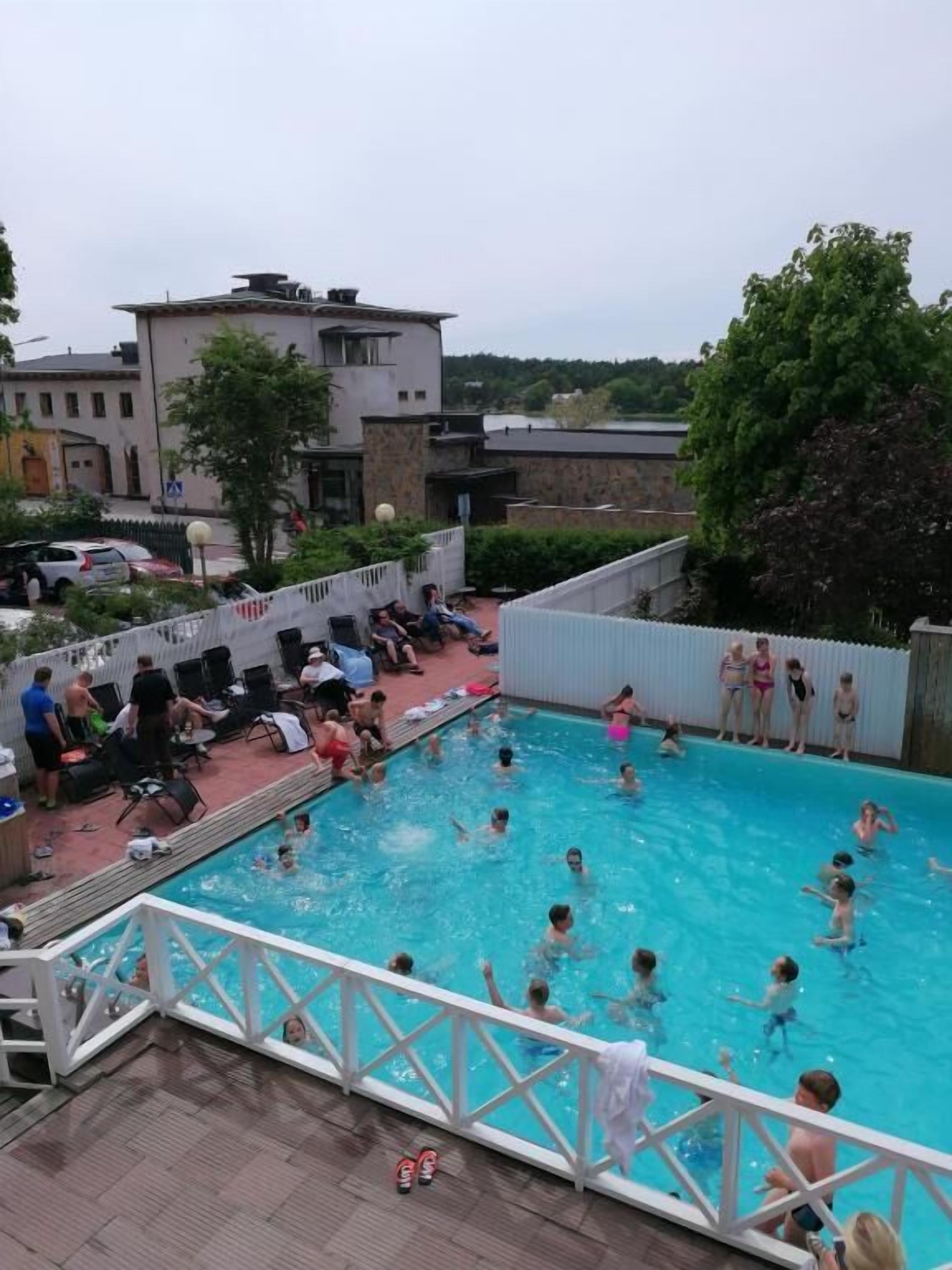 Ein Schwimmbad mit Personen, einem weißen Zaun und Gebäuden im Hintergrund.