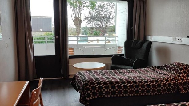 A hotel room with a bed, a chair, a small table, and a view of an outdoor area with a pool and trees. - Hotell Cikada (Mariehamn)