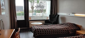 Ein Hotelzimmer mit Bett, Sessel, kleinem Tisch und Blick auf einen Außenbereich mit Pool und Bäumen.