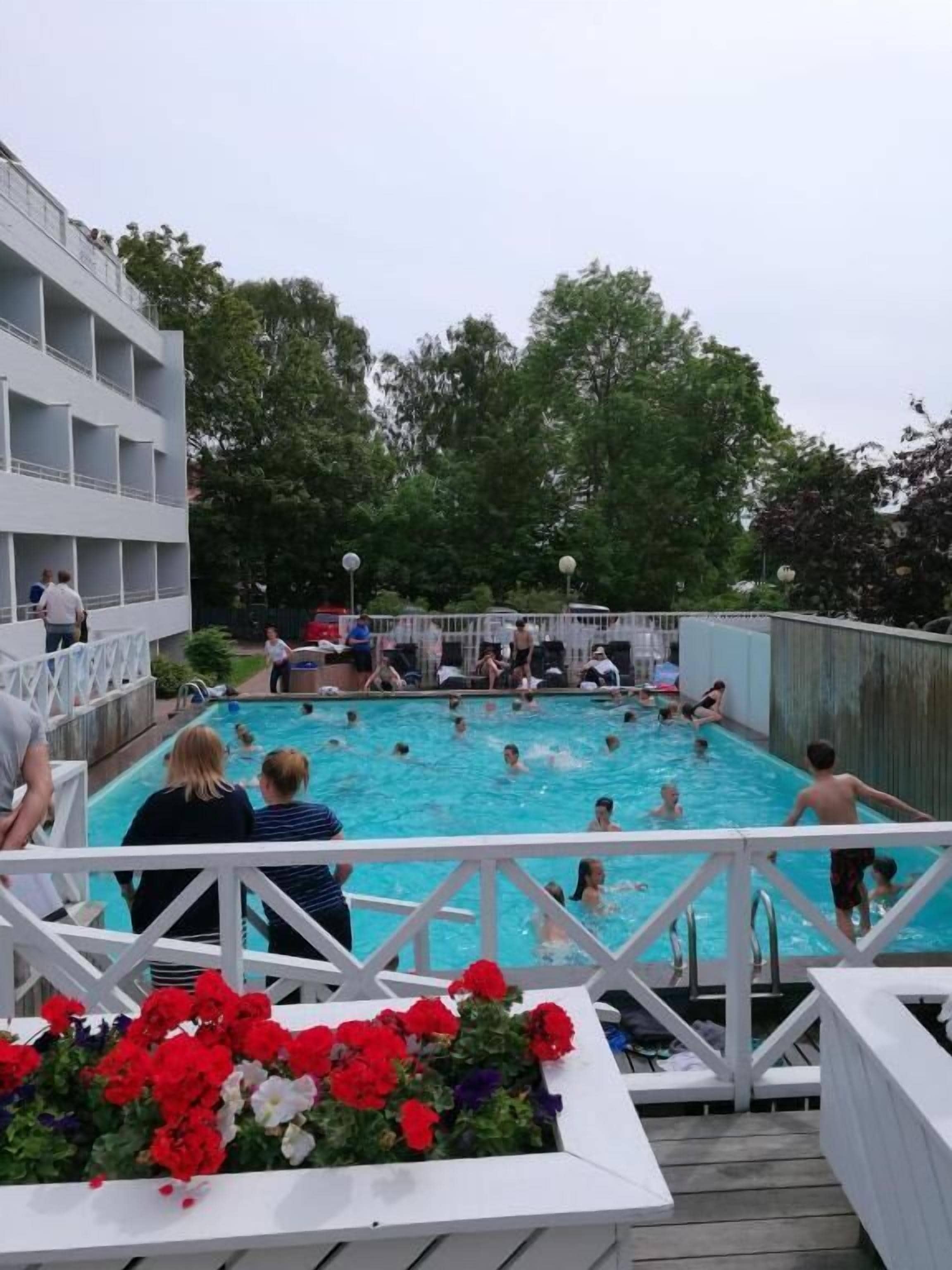 En swimmingpool med folk runt omkring, en vit byggnad med balkonger och en träveranda med blommor.