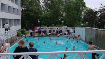 En swimmingpool med folk runt omkring, en vit byggnad med balkonger och en träveranda med blommor.