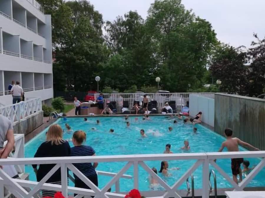 A swimming pool with people around it, a white building with balconies, and a wooden deck with flowers.