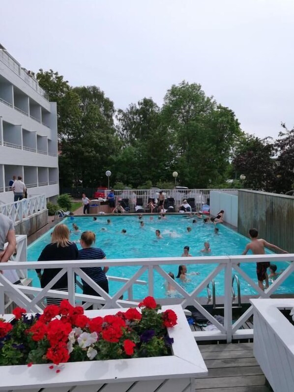 Una piscina con gente alrededor, un edificio blanco con balcones y una terraza de madera con flores.