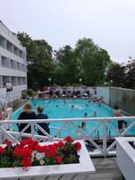 Una piscina con gente alrededor, un edificio blanco con balcones y una terraza de madera con flores.