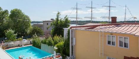 A swimming pool, a yellow building, and a view of the sea with sailing ships.