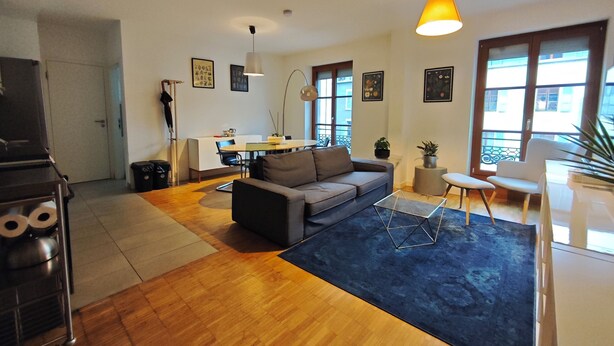 Ein modernes Wohnzimmer mit einer Couch, einem Couchtisch und einem blauen Teppich. Es gibt einen Essbereich mit Tisch und Stühlen sowie eine Küchenzeile mit Herd.
