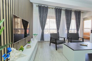 Ein modernes Zimmer mit an der Wand befestigtem Fernseher, zwei Sesseln, einem kleinen Tisch und einem Balkon mit Vorhängen.