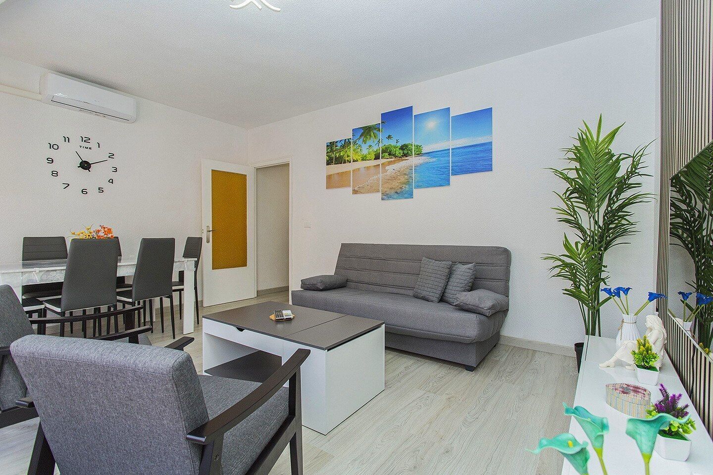 A modern living room with a grey sofa, a coffee table, and a dining area with chairs and a table. There is a wall clock and a series of landscape paintings.