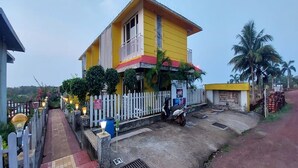 A yellow building with a balcony, a white fence, and a paved walkway. - White Sand Retreat (Guhagar)