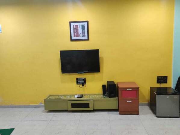 A room with a yellow wall, a flat-screen TV mounted above a green entertainment unit, a red cabinet, and a small grey refrigerator.