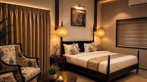 A hotel room with a bed, two nightstands, a chair, a round table, a potted plant, and a painting on the wall.