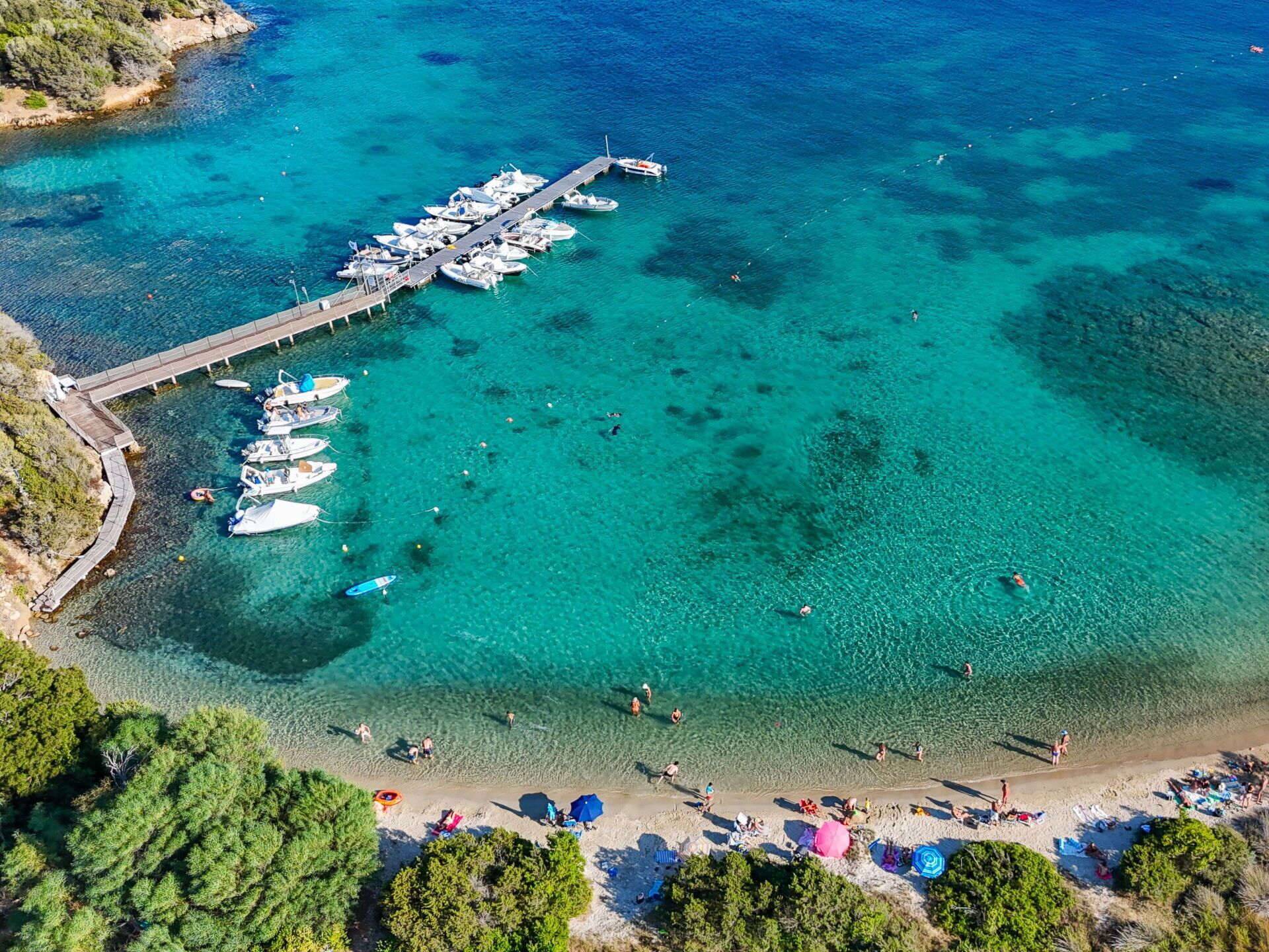 An aerial view of a coastal area with a marina, clear turquoise waters, and a sandy beach with umbrellas.