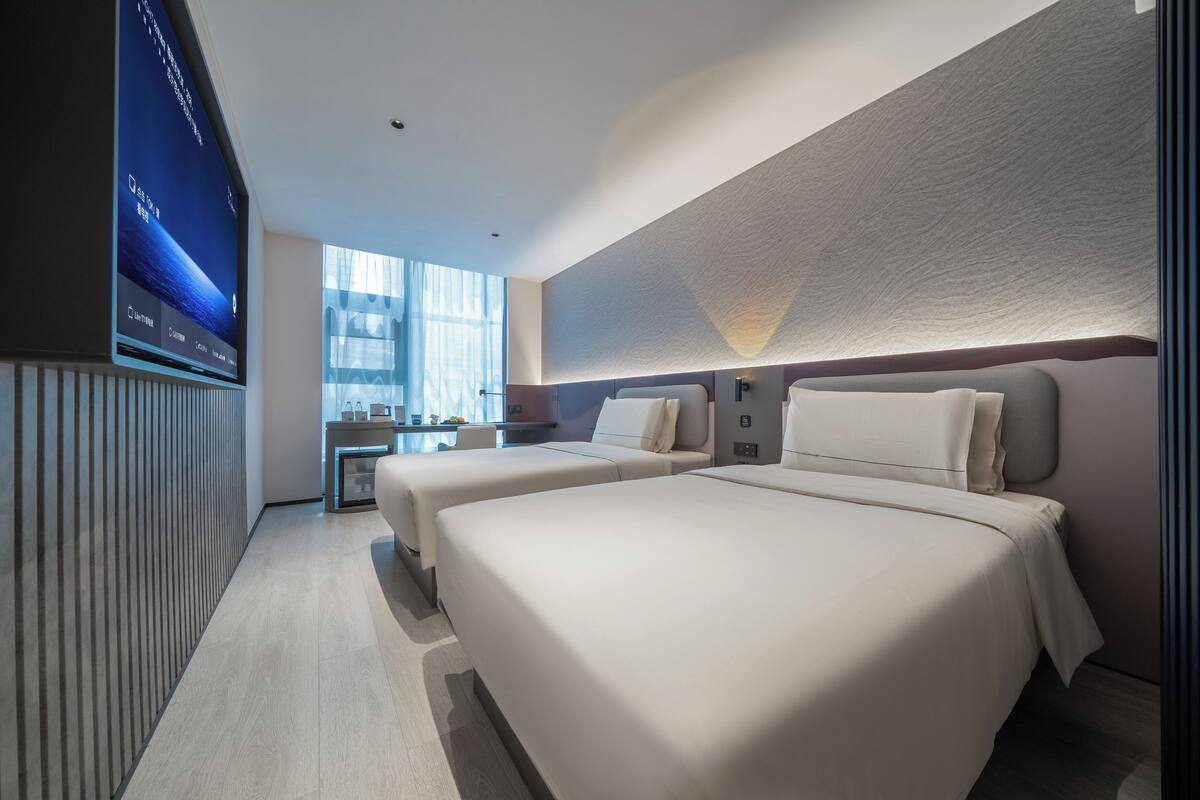 A modern hotel room with two beds, a TV, a desk, and a large window.