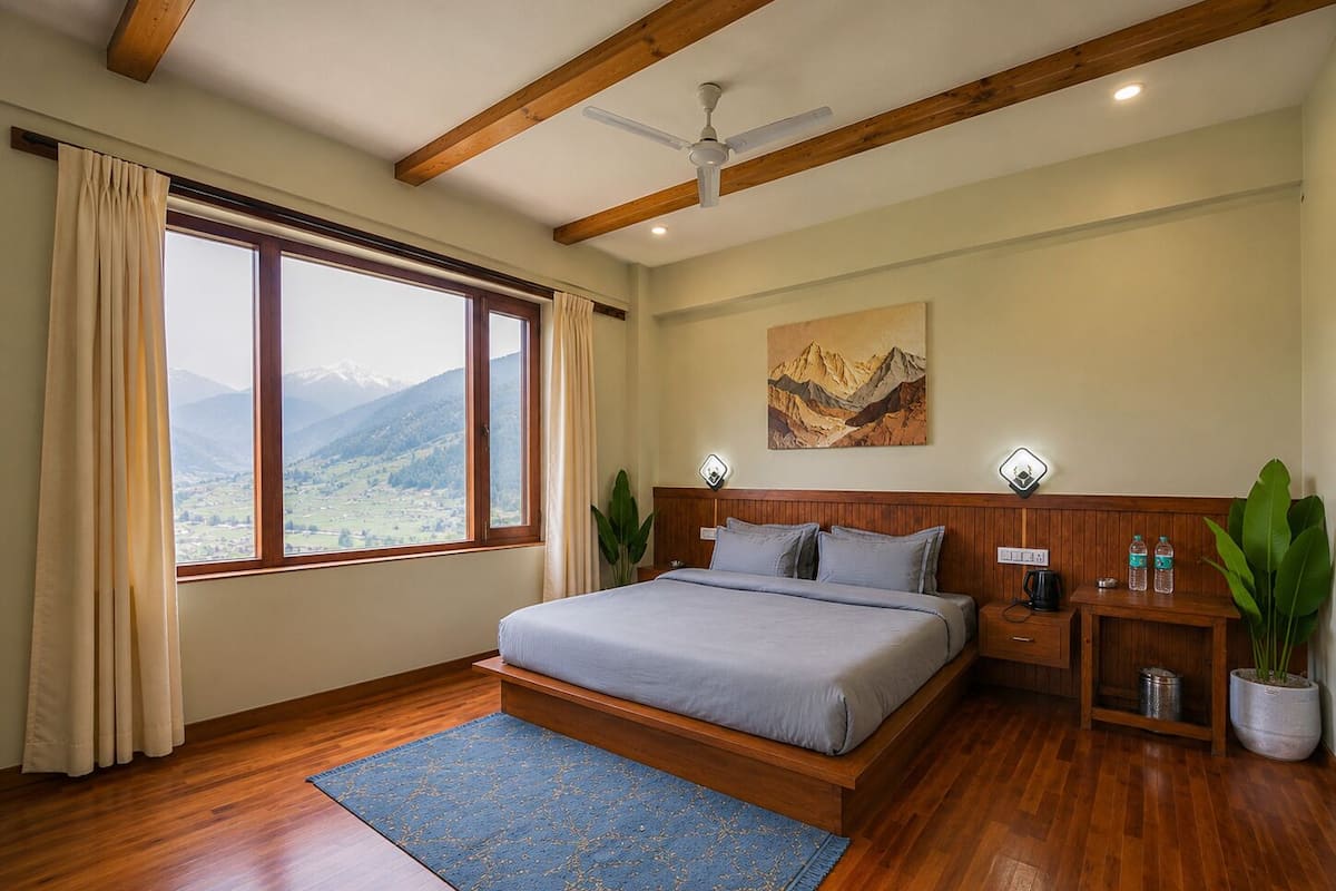 Un dormitorio con cama, vigas de madera, ventilador de techo, ventana con vista a la montaña, un cuadro, lámparas de noche y una mesita de noche con una planta.