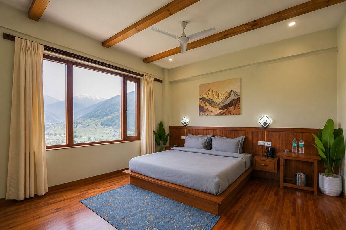A bedroom with a bed, wooden beams, a ceiling fan, a window with a mountain view, a painting, bedside lamps, and a nightstand with a plant.