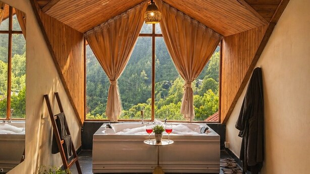 Um quarto com teto inclinado, vigas de madeira, uma janela grande com cortinas, um banco, uma mesa pequena com copos e um candeeiro suspenso.
