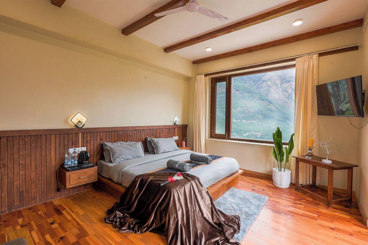 Un dormitorio con cabecera de madera, cama con ropa de cama gris, mesita de noche con cafetera y otra con una planta, y una ventana con vistas a la montaña.