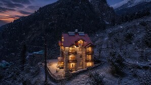 Uma cabana de registos de vários andares com telhado vermelho, rodeada por árvores e montanhas cobertas de neve.