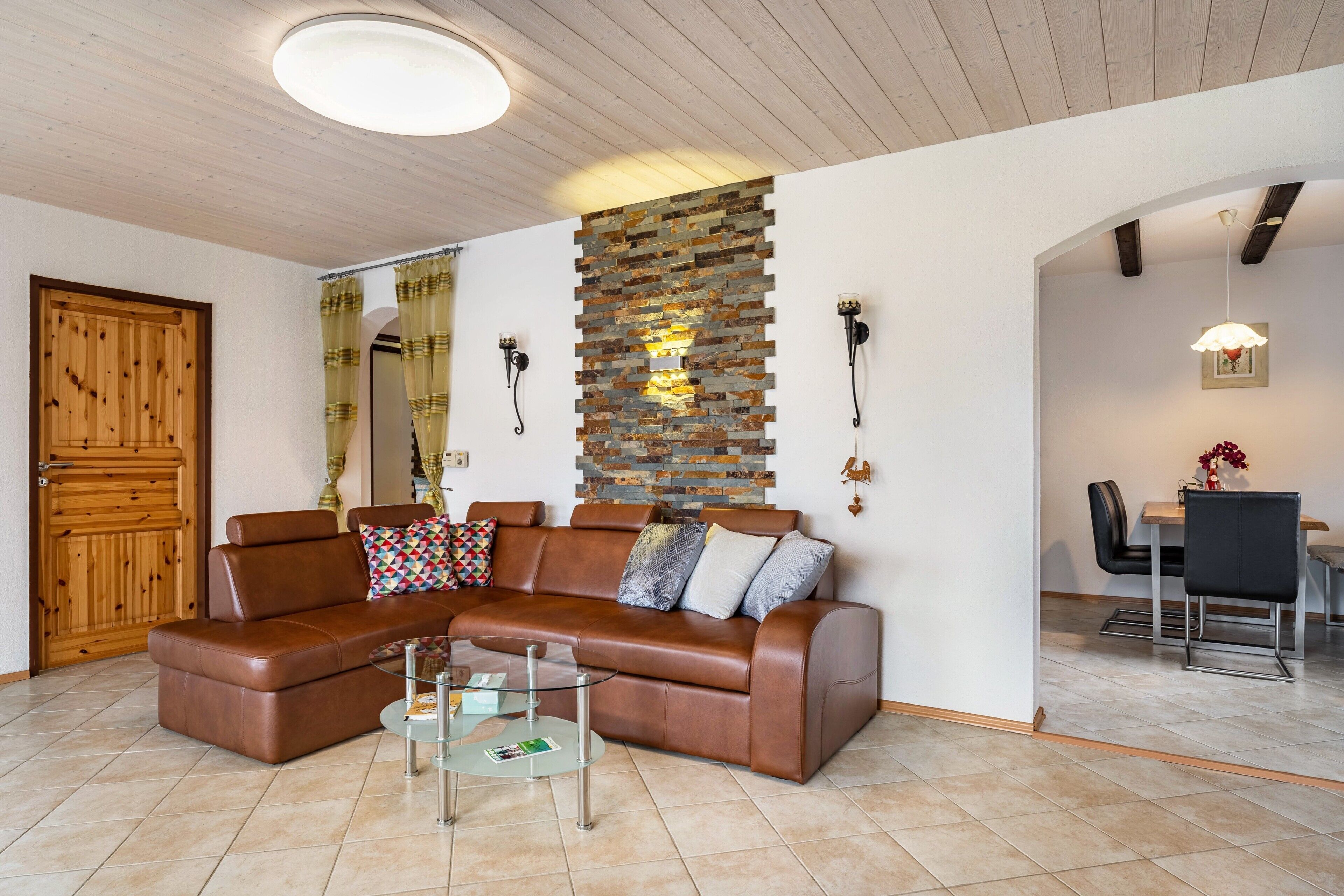 Ein Wohnzimmer mit braunem Ledersofa, einem Glastisch, einer Steinwand und einer Holztür.