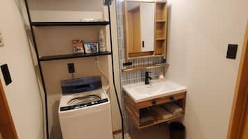 A compact bathroom with a washing machine, a sink, a mirror, and a shelf with toiletries.
