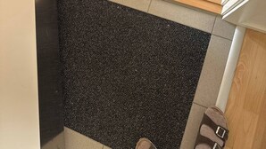 Rincón de una habitación con un tapete negro texturizado, suelo de madera y un marco de puerta blanco.