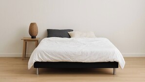 Una cama con edredón blanco, cabecera oscura y una mesita de noche de madera con una cesta tejida.